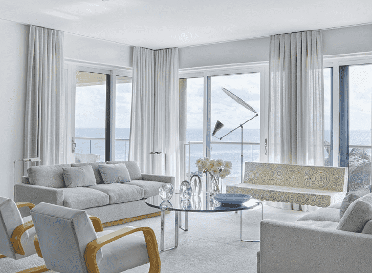 A living room with white furniture and a view of the ocean decorated with disposable curtains.